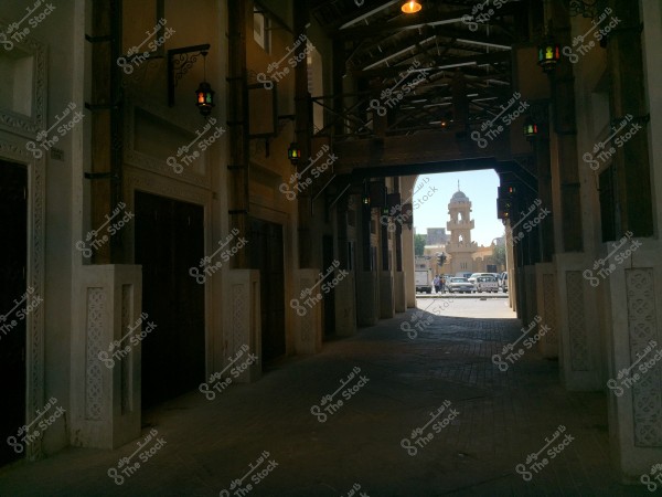 An image of a traditional alley with wooden ceilings and ornate windows on both sides, with colored glass lamps hanging from above. At the end of the passage, part of a city is visible, with a mosque tower in the background and a few cars and people.
