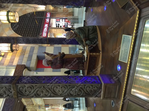 A statue of a man wearing a turban and traditional clothing seated at a table in an indoor area adorned with pillars and carvings, with a background showing part of a shopping center and two people. The scene reflects traditional architectural style and warm lighting.\r\n\r\n###