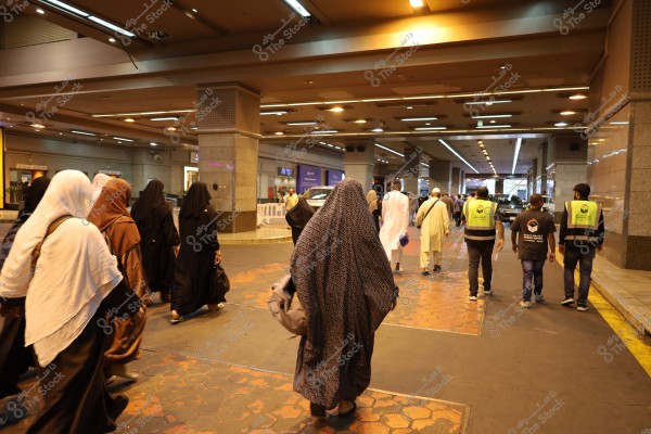 A group of people walking in an indoor area that appears to be a parking or transit station. Women are wearing abayas and hijabs in various colors like black and brown, while men are in white thobes and light-colored garments. Some individuals are wearing reflective vests with logos on the back. The architectural design suggests a modern, busy place with artificial lighting.
