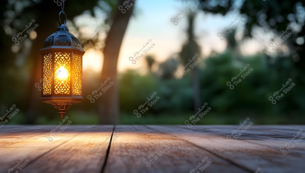 A decorative, illuminated Arabic lantern hangs against a blurred background of green trees at sunset. The glowing lantern, with its intricate details, creates a warm and tranquil atmosphere.