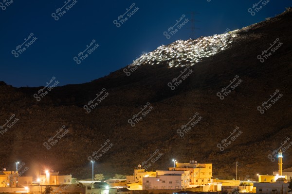 Night view of a village at the base of an illuminated hill. Houses are lit with warm lights scattered across the lower part of the image, with a mosque minaret illuminated on the right. The top of the hill is lit with white lights, and the sky in the background is dark.