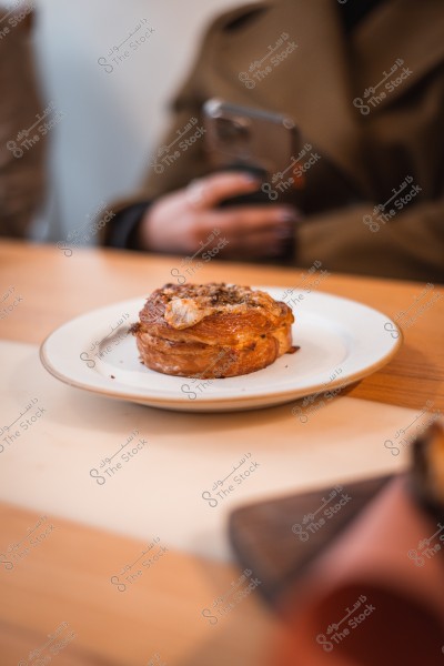 A piece of pastry on a white plate on a wooden table, with a person holding a phone in the background.