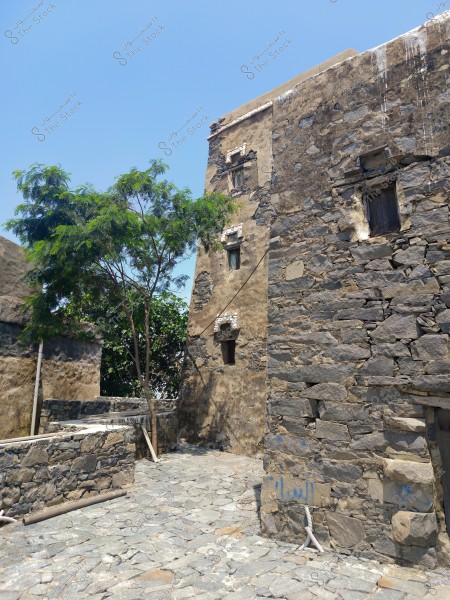 Image of a traditional stone building in an old village. The worn stone walls and small windows of multiple buildings are visible amidst a natural setting. A tree is clearly seen beside the building, and there is a stone-paved ground below. The sky is blue and clear, suggesting a sunny day.