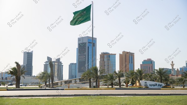 An image showing an urban scene of a Saudi city with a collection of modern buildings and palm trees. In the center, the Saudi Arabian flag is hoisted high. In the foreground, there is a green area and a road. There\'s also a sign that reads \"دوار التوحيد\".