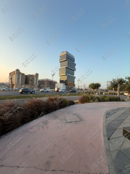 مبنى حديث بتصميم فريد يظهر مكوناً من عدة طوابق متكدسة فوق بعضها, يقع بجواره مباني تقليدية أخرى تحت سماء زرقاء صافية. يمكن رؤية السيارات على الطريق وأعشاب تزيينية على الرصيف القريب. الأفق يوحي بجو هادئ في وقت الغروب.

-