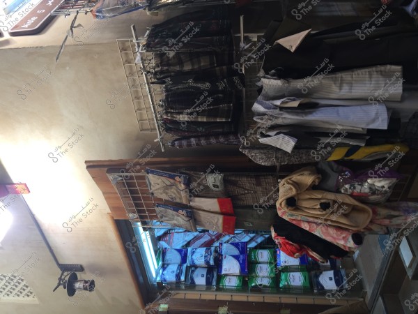 Image of a small clothing store displaying a variety of fabrics and hanging clothes. Shelves show shirts in different colors and patterns, along with pants ranging from formal to casual styles. There are several bags containing blankets or Ihram fabrics. The lighting is dim with a small sign indicating number 5 hanging on the wall.