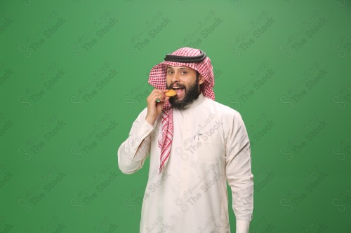 شاب سعودي على خلفية خضراء يرتدي الثوب و الشماغ، يقوم بعمل حركات بيده،يمسك الكعك و يأكله ، احتفالات العيد