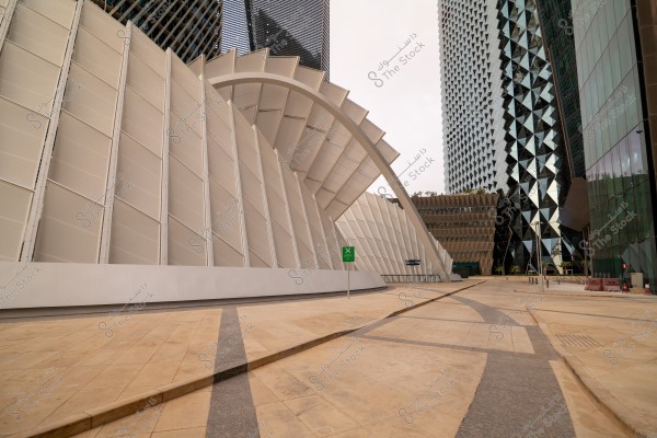 Image of modern buildings with glass facades and beautiful architectural geometric designs. A building with a curved facade designed with distinctive geometric shapes is visible, with a wide, empty tiled plaza in front. In the background, there are tall buildings with modern design and glossy glass walls reflecting the light.