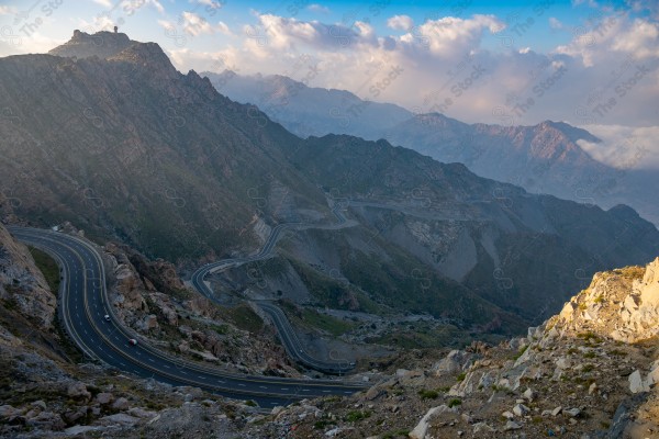 A snapshot of a series of towering rocky mountains and paved roads extending in the Al Hada Mountains in Taif, showing a cloudy sky during the day, mountain heights, Taif Mountains, mountain range