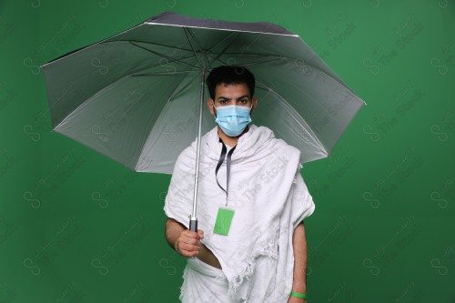 Portrait of a Saudi pilgrim on a green background wearing the Ihram to perform rituals while holding an umbrella in his hand to protect from the sun, wearing a mask