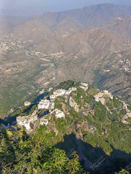 الصورة تُظهر منظرًا جويًا لقرية تقع في منطقة جبلية خضراء. البيوت مبنية على سفح الجبل, وهي ذات لون أبيض وتحيط بها طرق ملتوية. الجبال المحيطة ذات قمم غير منتظمة وتنحدر بلطف نحو الأفق. النباتات الخضراء تغطي أجزاءً من التلال, مما يُضفي على المكان مظهرًا خصبًا.