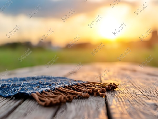 A blue patterned hat with brown fringes on a wooden surface in an outdoor setting with a sunset in the background. The sky is colored with shades of yellow and orange, creating a peaceful gradient, and there\'s a blurred greenery in the background that adds a sense of tranquility and nature.