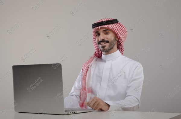 A portrait of a man sitting in front of a silver laptop. The man is wearing traditional Saudi attire, a white thobe with a red and white checkered shemagh and a black agal. The background is plain and light gray.