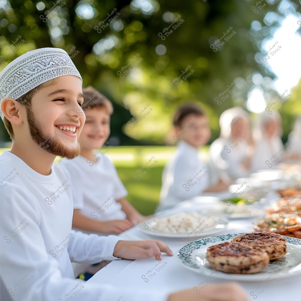 يظهر في الصورة مجموعة من الأطفال يجلسون حول طاولة في الهواء الطلق وهم يرتدون ملابس بيضاء تقليدية. الأولاد يبدون مبتسمين أثناء استمتاعهم بالطعام الذي يبدو متنوعًا, بما في ذلك أطباق من الأرز واللحوم. البيئة المحيطة خضراء ومشرقة, مما يشير إلى أنها نهار في حديقة أو في مكان طبيعي.