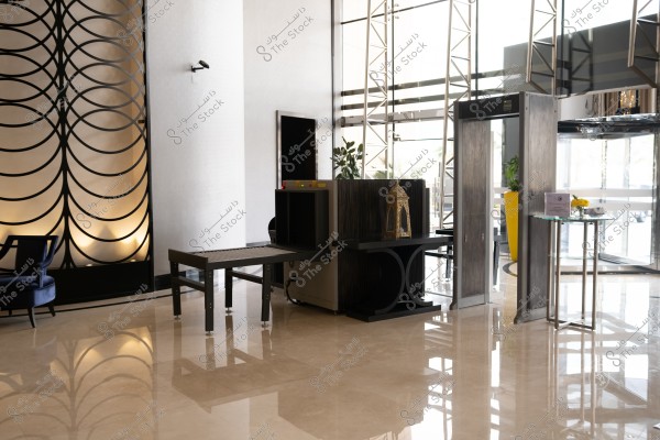 يظهر في الصورة مدخل فندق مع جهاز كشف المعادن وطاولة صغيرة تحمل إناء للزهور. الجدران مزينة بنمط هندسي وفوانيس ذهبية, وتنعكس الإضاءة الخافتة على الأرضية الرخامية. يظهر في الخلف كرسي مفرد وزرع في أصيص لونه أصفر.