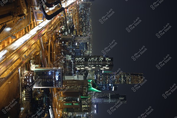 A night-time image of a modern architectural cityscape, featuring tall skyscrapers illuminated against the night sky, and a busy main street filled with traffic, showcasing streams of headlights and taillights. The city lights add vibrancy and highlight the beauty of the modern architecture.