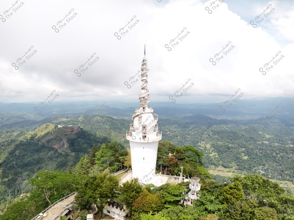 Image of a tall white tower with a spiral design at the top, situated in the middle of a lush green mountainous area with dense forests surrounding it. Clouds cover the sky in the distant horizon, adding a sense of mystery and beauty. Roads and terraces near the tower indicate human activity in the area.