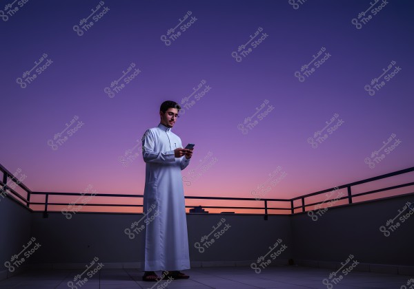 A man standing on a balcony at sunset, wearing a white thobe and using a mobile phone. The sky is beautifully colored with shades of purple and orange, and there\'s a metal railing around the balcony.