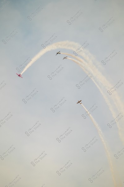 Four small planes are performing in the sky, leaving behind trails of white smoke. The left plane is red, and they are all following curved paths in the air.