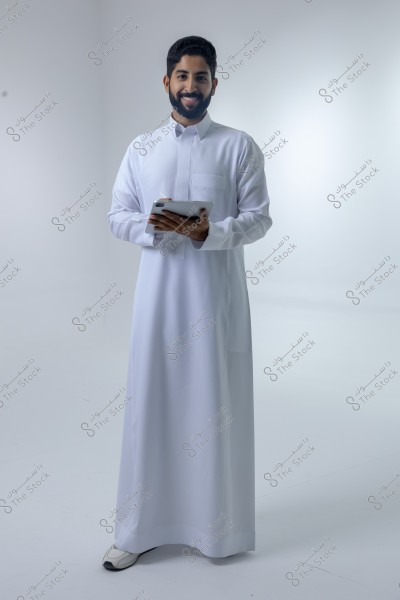 A portrait of a man wearing a traditional white thobe and smiling at the camera. He is holding a tablet and standing in a studio with a white background. The thobe suggests the man may be from the Gulf region.