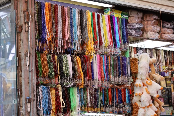 An image of a shop in a market displaying a variety of colorful prayer beads hanging on the wall. The beads come in multiple colors such as red, brown, green, blue, and more. Next to the beads are some plush toys shaped like camels, with white bodies and orange details. The shelves in the background show slippers wrapped in plastic and other products.