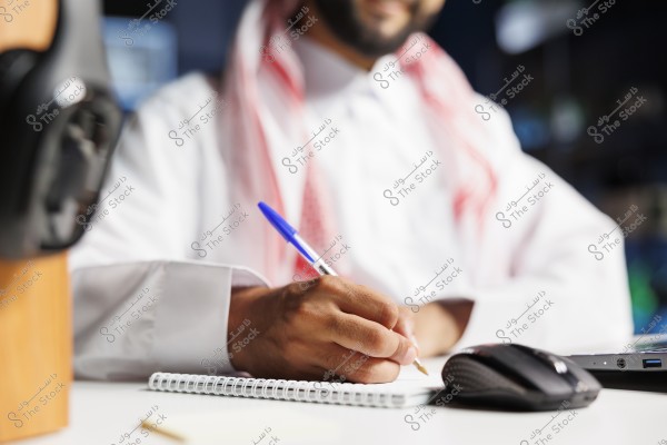 The image shows a man wearing a white thobe and a traditional red keffiyeh, sitting at a desk. He is focused on writing in a notebook with a blue pen. A computer mouse and part of a laptop are visible beside him. The setting suggests a work or study environment indoors.