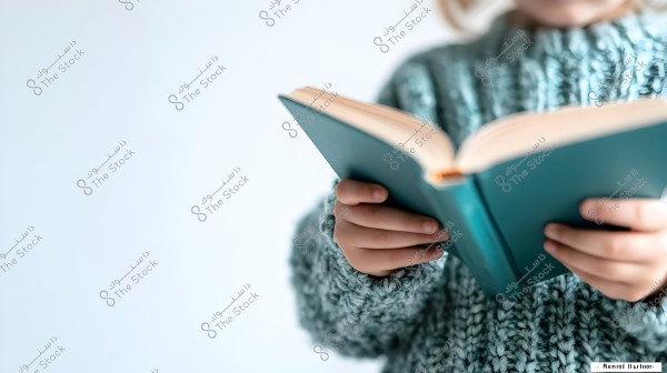 An image of a child wearing a knitted sweater, reading a predominantly blue book. The background is white, with a focus on the hands and the book.