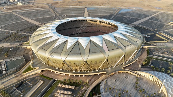 An aerial view of a modern stadium with a petal-like white roof, surrounded by a vast open area and parking lots. The stadium features unique architectural design with golden geometric shapes adorning the exterior walls. Sunlight reflects off the roof and the surrounding area.