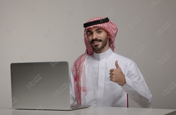 An image of a man sitting in front of a laptop on a table, giving a thumbs-up gesture. He is wearing traditional Saudi attire, including a white thobe and a red checkered shemagh. The background is plain and light gray.