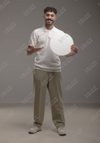 A man standing in front of a gray background, wearing a white polo shirt, beige trousers, and sneakers. He is holding a white circular board in his right hand and is smiling at the camera. The lighting is even and focuses on the person in the image.