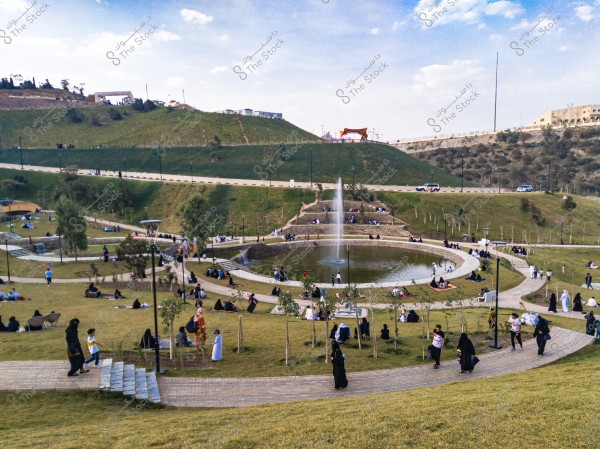 The image depicts a public park on a sunny day, with many people gathered to enjoy the outdoors. A large circular fountain is shown in the center, surrounded by green spaces and gentle hills. People are sitting on the grass and walking on pathways. Some are wearing traditional Saudi attire.