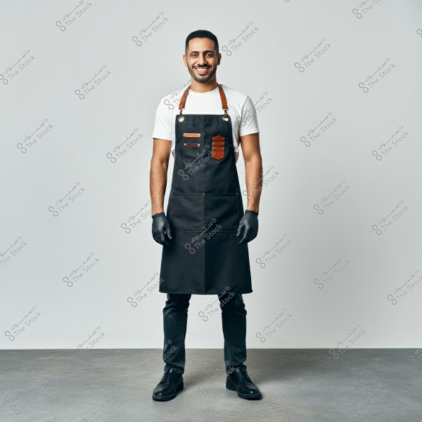 A photo of a man standing, wearing a black apron with leather details and a white shirt. The man is wearing black gloves, dark pants, and black shoes. The image appears to be taken in a studio with a simple gray background. The attire suggests a professional setting such as cooking or barbering.\r\n\r\n###