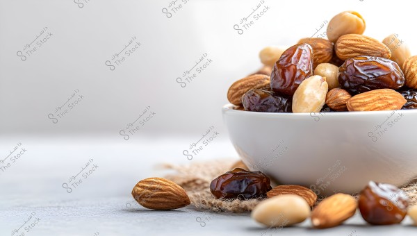 An image showing a white bowl filled with dates and almonds on a gray surface. The dates appear glossy and dark brown, alongside brown almonds and white almonds. Some almonds and dates are scattered around the bowl. The background is soft, giving a calming impression.