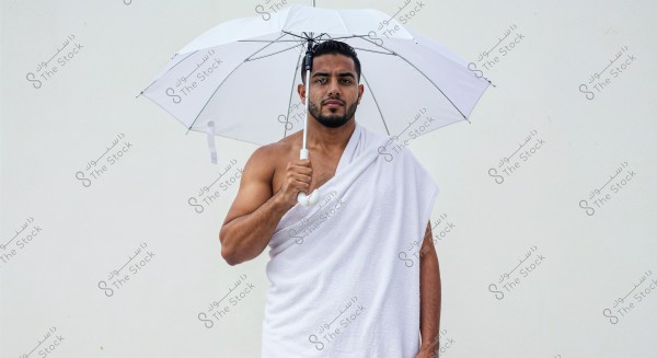 Image of a man standing against a white background, wearing the Ihram attire associated with Hajj or Umrah rituals. He is draped in a white cloth from his shoulders to below his knees. He holds an open white umbrella for sun protection.