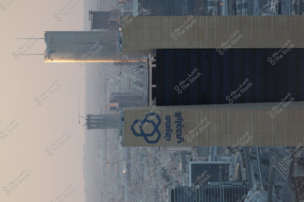 Image of two high-rise buildings in a modern city at sunset. One of the buildings has a large sign written in Arabic. The background features a cluster of towers and tall buildings, alongside a network of roads and bridges. The sunlight fades on the horizon, casting warm shadows across the scene.