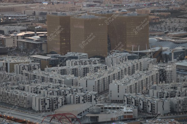 منظر جوي لمدينة الرياض يظهر مجموعة من المباني السكنية الحديثة ذات التصميم المعماري الفريد, مع أبراج ذهبية شاهقة تظهر في الخلفية. الأبراج محاطة بمجمعات تجارية ومرافق عامة, وتبرز مساحات خضراء وعدة شوارع تربط المنطقة ببعضها.