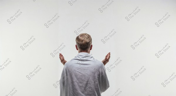 An image of a man standing with his back to the camera, raising his hands to the sides in an unclear position. The man is wearing a white robe and stands in front of a plain white background. The lighting is even, contributing to a calm and contemplative atmosphere.