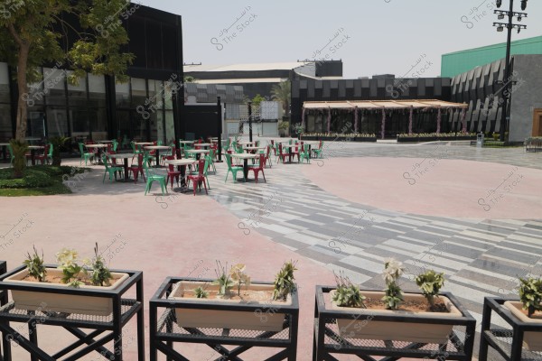 The image shows an open outdoor plaza with several tables and chairs colored in green and red. In the foreground, there are planter boxes with flowers, and the front facade is a modern building with large glass windows. In the background, there are other structures and green plants, with fabric canopies over some areas.
