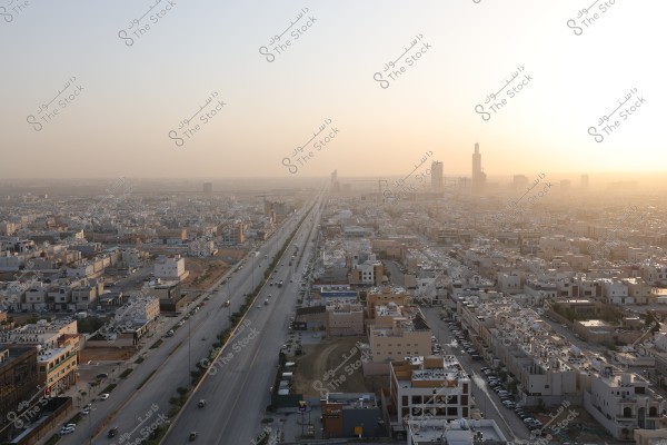An aerial view of a modern city during sunset on the horizon. Residential and commercial buildings are closely packed, with a wide street running through the center of the image. Some distant towers are visible at the city\'s edge, with clouds painted with sunset colors filling the sky.