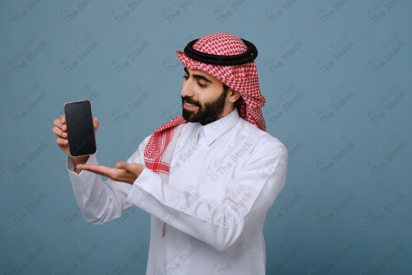 بورتريه شاب سعودي يرتدي الزي السعودي يشير إلى هاتفه ويتفاعل معه، وتظهر على ملامحه الدهشة ، على خلفية سماويه