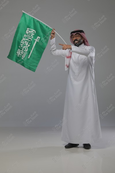 A man wearing traditional Saudi Arabian white thobe and a red and white checkered shemagh, holding the flag of Saudi Arabia. The flag is green with a sword and the words \"There is no god but Allah, Muhammad is the messenger of Allah\". The man stands against a grey background.