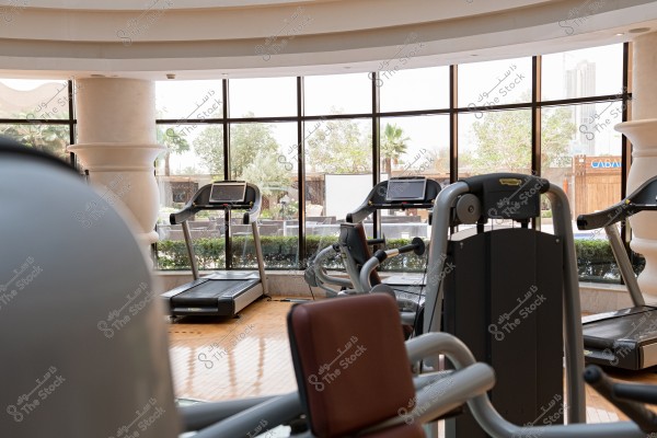 تُظهر الصورة الداخل الأنيق لصالة رياضية حديثة مع نوافذ كبيرة تسمح بدخول الضوء الطبيعي. توجد عدة آلات جري وتمارين رياضية أخرى موزعة على أرضية خشبية, في وسط مساحة مفتوحة وواسعة. خلف النوافذ, يمكن رؤية أشجار النخيل وشجيرات خضراء, مما يعطي شعورًا بالاندماج مع الطبيعة.