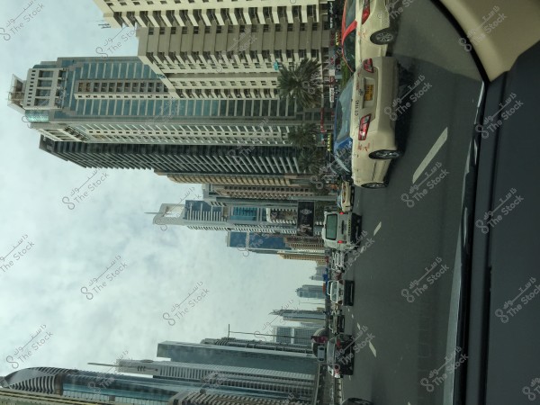 The image depicts a modern cityscape with tall skyscrapers on either side of a wide road. Many cars are driving along the street that runs between the buildings. The sky is overcast with clouds, allowing some light to pass through. Palm trees line the sidewalk.