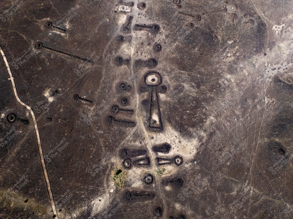 An aerial view of a desert area showcasing ancient and varied stone formations standing out against the brown earth. The formations include circular and rectangular sites arranged in a specific order, possibly part of mysterious historical structures or archaeological sites in a remote area.