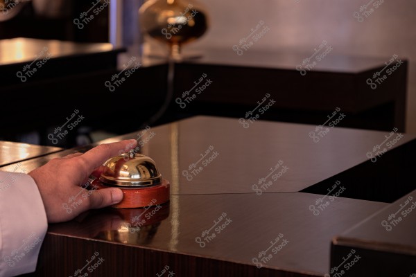 يد شخص يرتدي قميصاً أبيض وهو يضغط على جرس استقبال معدني ذهبي موضوع على طاولة خشبية داكنة اللون, يشير إلى بيئة فندق أو مكتب استقبال, مع ظلال دافئة ومصباح ذهبي في الخلفية.