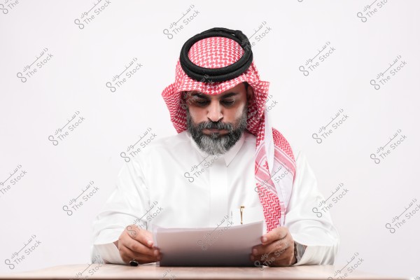رجل يرتدي الثوب والغترة والعقال يقرأ وثيقة جالسًا على طاولة.