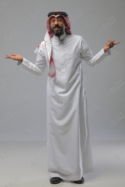 An image of a man standing and smiling, wearing a traditional white thobe and a red and white checkered headscarf with a black agal. The man has his hands raised as if talking or explaining something. The background is a neutral gray.