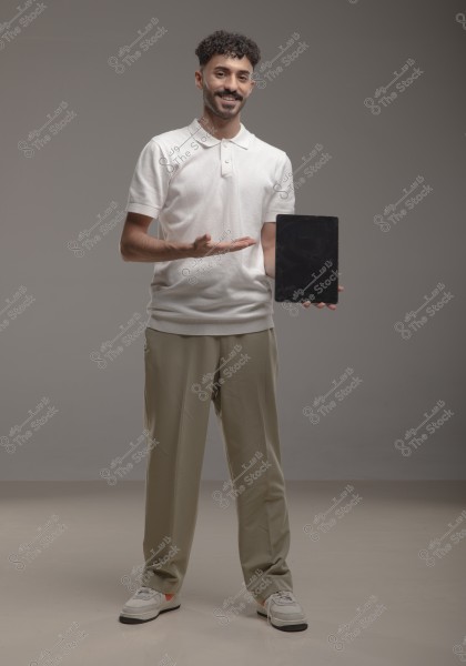 A portrait of a man standing in a simple studio, wearing a white polo shirt, beige pants, and white sneakers. He appears to be presenting, gesturing with one hand toward a black tablet he holds in the other. The background is gray, giving the impression of a professional shoot.