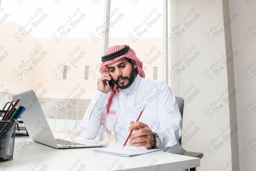 رجل سعودي يرتدي الزي السعودي التقليدي يعمل في مكتب بواجهة زجاجية ويستخدم قلم رصاص لتدوين الملاحظات