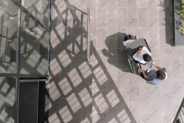 A person pushing another individual in a wheelchair beside a glass building.